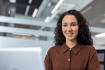 Young woman with curly hair smiling and looking at a laptop screen, actively working and...