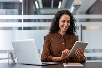 Professional businesswoman with curly hair smiling while using a tablet at a modern desk, multitasking with laptop, showcasing confident remote work, digital connectivity and success