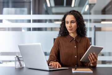 Young professional businesswoman sitting at a desk and simultaneously working on a laptop computer and a digital tablet, effectively managing tasks in a modern office environment