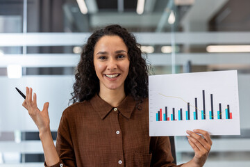 Confident businesswoman presenting a financial data report chart, holding a pen and smiling, showcasing success, growth, and effective communication in a modern office environment