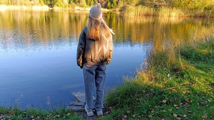 A girl in a warm jacket and a knitted hat stands on the shore of a lake.