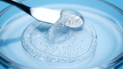 A stainless steel spatula spreads a gel substance in a clear glass dish on a light blue backdrop
