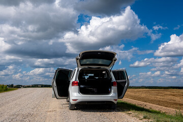 A white car with open doors and trunk stands on a dirt road in a field against the backdrop of a...