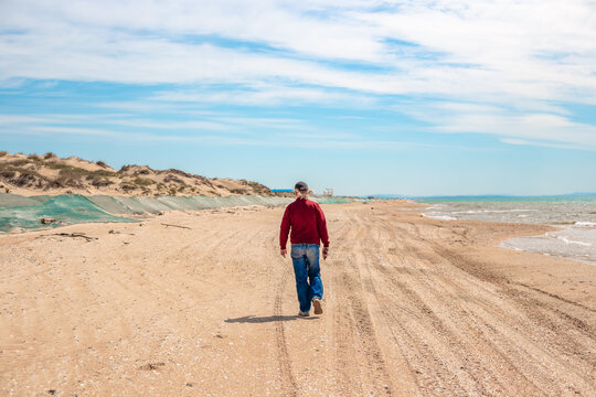 A man walks along a sandy beach on a sunny day. Along the sea, a sand embankment is covered with a net to catch fuel oil