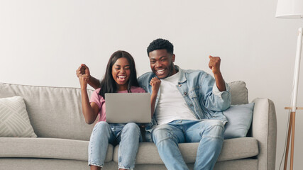 Great Job, Win And Luck Concept. Excited black couple using pc laptop, cheering looking at screen, celebrating victory. Emotional happy guy and lady shaking clenched fists, sitting on sofa, free space