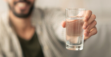 A man holds a transparent glass filled with water, smiling warmly as he offers it to someone. The background is softly blurred, creating a relaxed atmosphere.