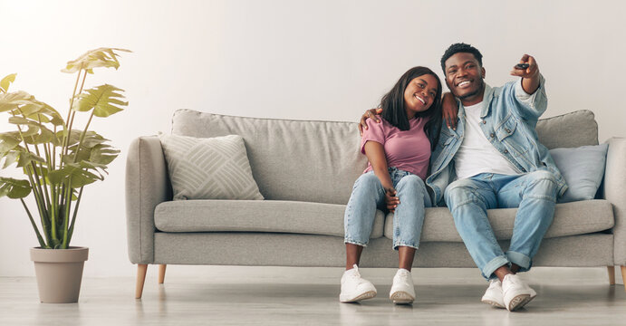 Weekend Leisure. Happy relaxed millennial black guy and lady watching tv in living room interior, holding remote control and hugging. Smiling couple enjoying film and movie evening together - Powered by Adobe