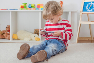 Cute little child using a smartphone while sitting on the floor at home. Digital device and screen time addiction, technology in the hands of children concept.