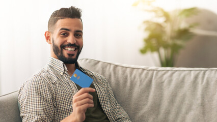 A man sits on a comfortable gray sofa, smiling broadly while holding a blue credit card. Sunlight streams through the window, creating a warm and inviting atmosphere in the room.