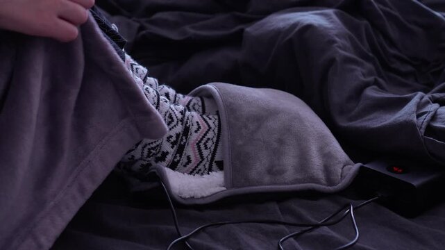 A woman in a bathrobe and patterned woolen socks warms her feet in an electric soft heating pad from a power bank on the bed. Cold season, warm, no heating. Cold colors