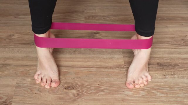 Caucasian woman trains legs and feet with elastic band. Lifts up and spreads legs. Exercises for rehabilitation of joints and ligaments. Home workout