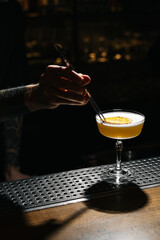 Bartender garnishing a yellow cocktail with a slice of dried orange using tweezers at a dimly lit bar. Elegant mixology, luxury drink and nightlife concept.