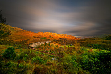 Golden sunset over the famous Glenfinnan Viaduct in the Scottish Highlands, where warm light...