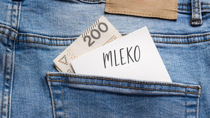 White card with a handwritten inscription "Mleko", put into the pocket of blue pants jeasnow, next to Polish banknotes PLN (selective focus)