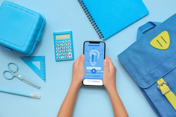 Child's hands with mobile phone and school stationery on blue background, top view. Audiobook concept