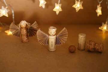 DIY Christmas angel figurines made from wine corks and paper wings with drawn faces and twine hair on brown background with glowing star lights. Christmas and winter holiday craft concept. light bokeh