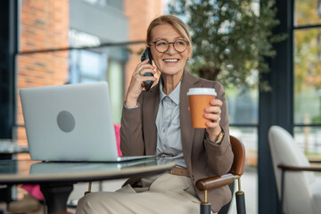 Happy mature businesswoman talking on smartphone holding coffee