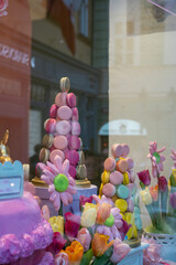 Colorful pastel macarons rise into towers beside bright flowers, creating a whimsical storefront display that evokes sweetness, celebration, and playful dessert art.