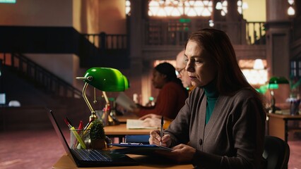 Intellectual mature woman transcribing academic text from laptop at the university, sitting in vintage study room and completing online documents. Education through formal learning.