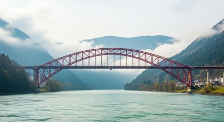 Obraz premium Red arch bridge spans a greenish-blue river, cutting through a misty mountain valley. Distant snow-capped peaks tower as calm water mirrors the sky and steel girders, creating a tranquil panorama now