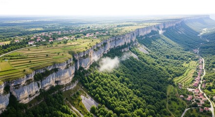 Obraz premium A dramatic canyon with towering limestone cliffs, emerald forests, and terraced fields along a sunlit plateau. A mist plume drifts from the cliff, while a winding road and a village echo along the rim