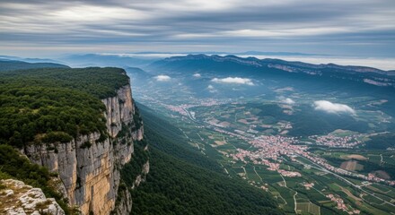 Fototapeta premium Cliffside panorama sheer rock faces, a valley below with patchwork fields, a winding road, and a distant village; mist drifts over treetops as a broad sky seals the landscape in cool light. With calm