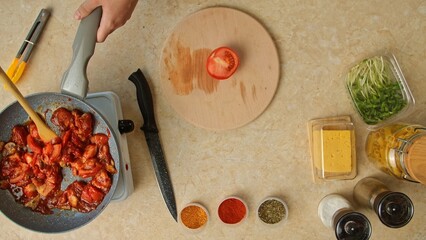 A top shot of the kitchen area with fresh vegetables, spices, cheese, and pasta, emphasizing the process of cooking. A woman is frying pieces of chicken with tomato sauce in a pan.