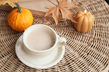 Cup of pumpkin coffee with candle, book and autumn leaf on basket in room, closeup
