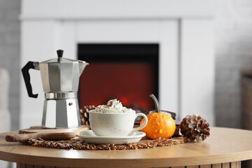 Cup of pumpkin latte with fir cone and coffee maker on table in room, closeup