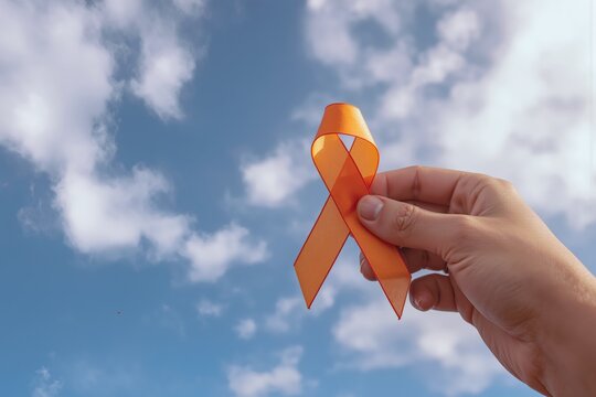 Hand holding an orange awareness ribbon against a blue sky, symbolizing support for health campaigns and causes