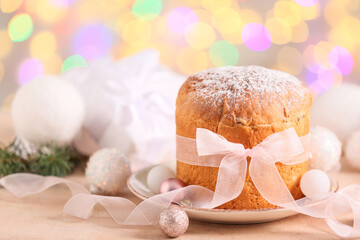 Tasty Panettone with bow and Christmas balls on table against blurred lights, closeup