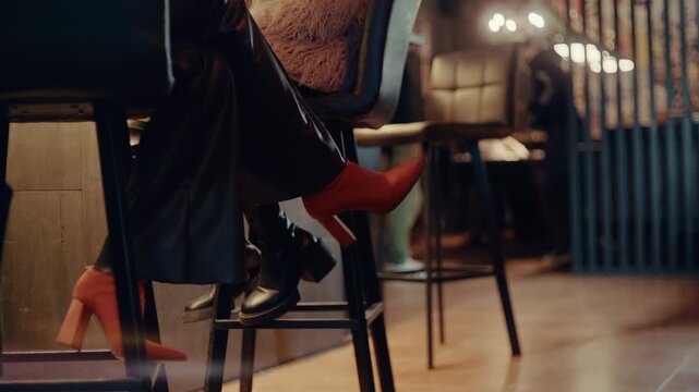 Close-up shot of two friends legs in red high heels sitting on bar stools at a modern night bar under warm ambient light.