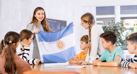 Fototapeta premium Group of preteen schoolchildren attentively watching pedagogue describing Argentina flag in schoolroom