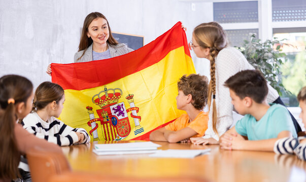 Decent teacher showing Spain flag to group of preteen schoolchildren in classroom during lesson
