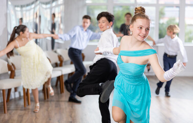 Obraz premium Cheerful girl in festive blue dress learning vigorous jive moves with partner during group lesson in choreography studio. Teenagers hobbies concept..