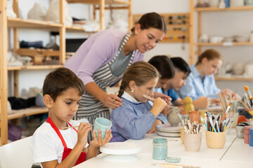 Focused boy paints a mug made of clay at a workshop. Boy at a children's master class on ceramics next to a teacher