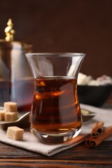 Tasty Turkish tea in glass cup, brown sugar, cinnamon, sweets and teapot on wooden table against color background, closeup