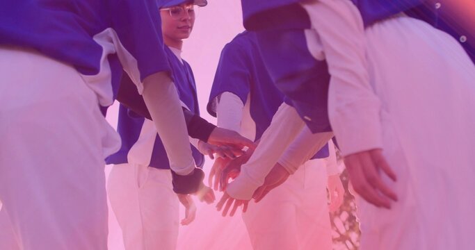 Six female baseball players placing hands in huddle on grass field in sunlight, with blue jerseys - Powered by Adobe
