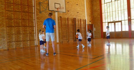Coaching coach holding orange ball guiding players dribbling in school gym, with climbing frames
