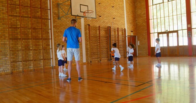Coaching coach holding orange ball guiding players dribbling in school gym, with climbing frames - Powered by Adobe