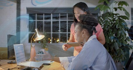 Two women wearing coral top collaborating over code in office at night, with laptop and tablet