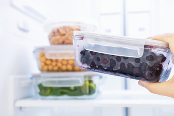 A hand holds a glass box with blueberries against the background of an open refrigerator, close-up.