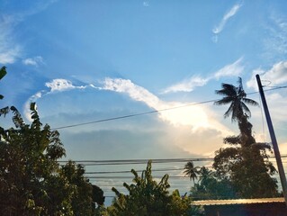A view of the sky with a large, dramatic cloud formation, with the sun's rays shining through.&nbsp;The foreground features silhouettes of tropical trees, including a palm tree, and power lines.