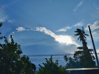 A view of the sky with a large, dramatic cloud formation, with the sun's rays shining through.&nbsp;The foreground features silhouettes of tropical trees, including a palm tree, and power lines