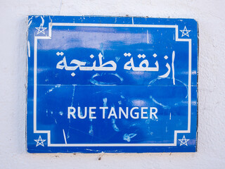 A weathered blue street sign displays "RUE TANGER" in white French and Arabic script, featuring Moroccan stars on a textured white wall.