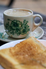 A classic Kopitiam breakfast of hot Kopi Susu (coffee with condensed milk) in a traditional floral porcelain cup, with a slice of Kaya Toast slightly blurred in the foreground