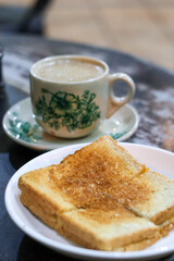 A classic Kopitiam breakfast of hot Kopi Susu (coffee with condensed milk) in a traditional floral porcelain cup, with a slice of Kaya Toast slightly blurred in the background.