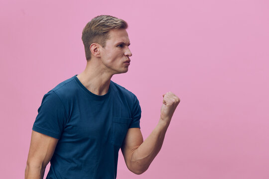 angry young caucasian man clenched fist frustrated emotion blue t-shirt side view isolated pink background mental health gadgets concept