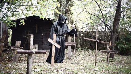 Grim reaper death in an old cemetery graveyard holding a scythe with wood crosses during gloomy rain. Halloween classic horror. Slider shot in slow motion. - Powered by Adobe