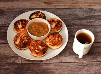 Fried cheesecakes with raisins on a large plate with apple jam on a wooden table, a cup of hot tea nearby.